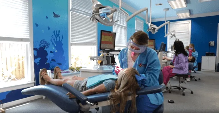 Child receiving dental care in a pediatric dentistry office, surrounded by colorful decor and dental equipment, with a dental assistant in a blue coat providing treatment.