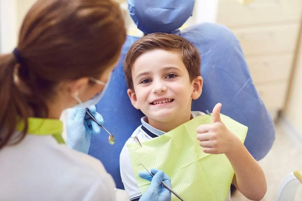 Smiling boy in dentist chair giving thumbs up, pediatric dentistry setting, highlighting positive dental experience related to tongue-tie treatment.