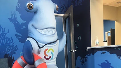 Shark mascot in dental office with colorful branding, promoting a friendly atmosphere for pediatric patients at Smiles From Us locations.