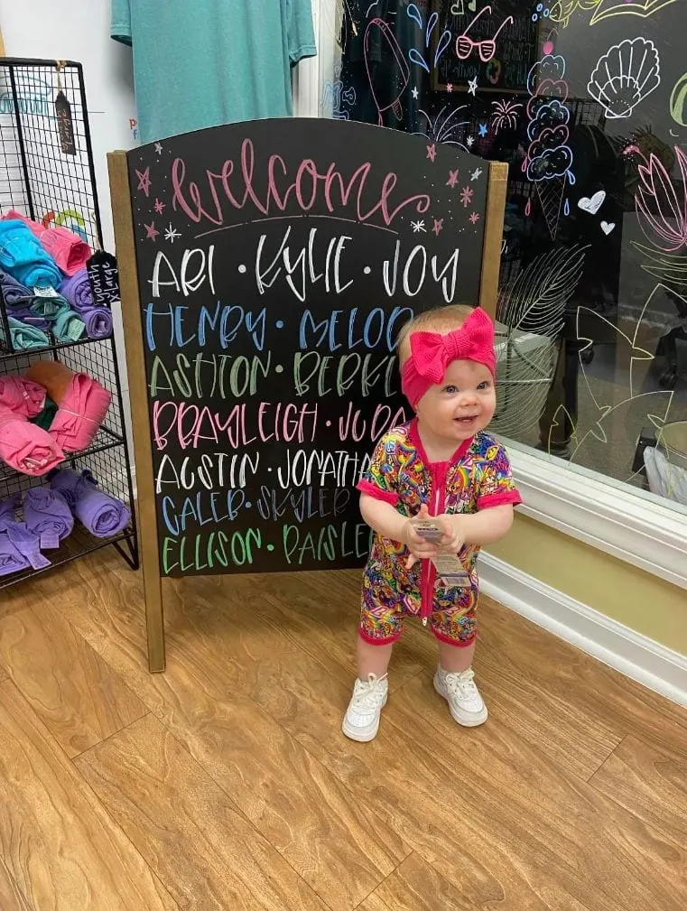 Toddler in colorful outfit standing in front of a welcome sign with names, promoting a friendly atmosphere for children, relevant to pediatric dental services.